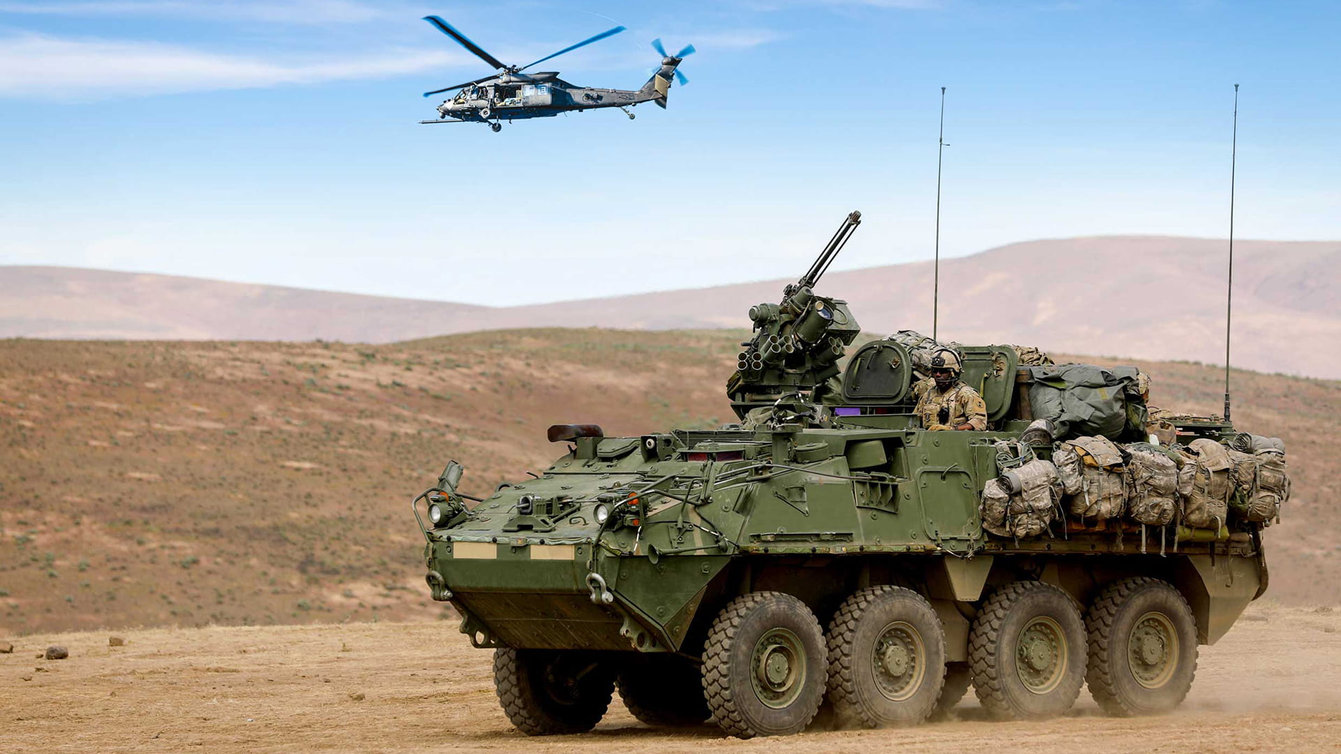 A U.S. Army Stryker tank in the desert. A Black Hawk helicopter can be seen in the sky.