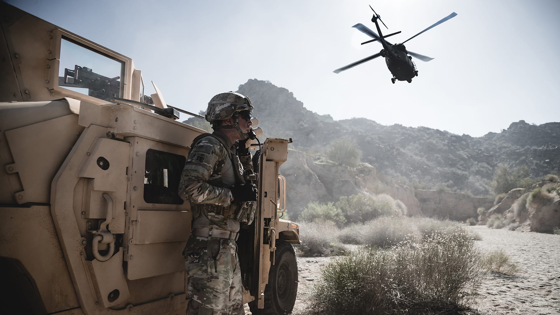 A solider stands next to a military Humvee. A helicopter flies overhead.