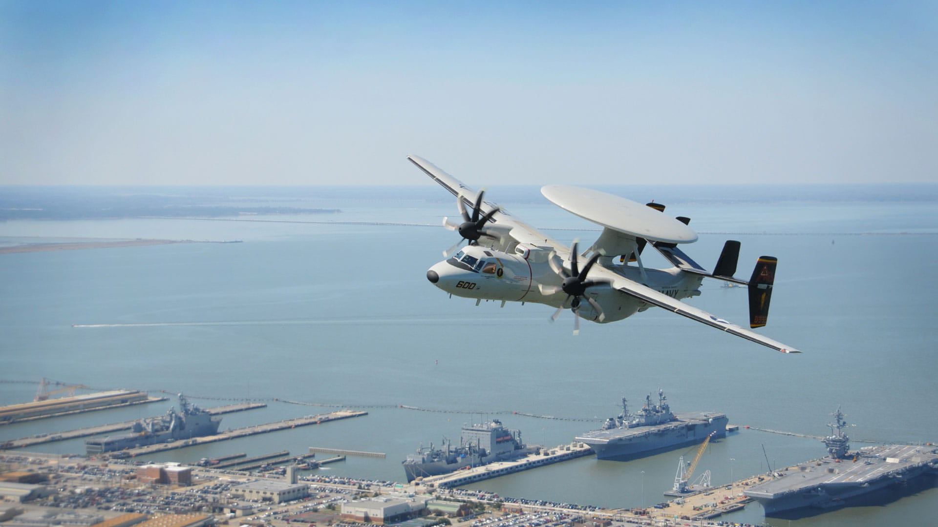 An E-2D in the air with Naval vessels visible in the background
