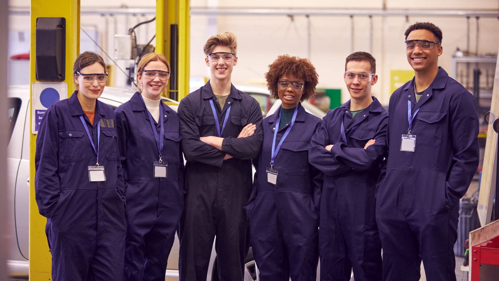 A group of six young apprentices, all wearing blue jumpsuits, stand in a workshop smiling at the camera.