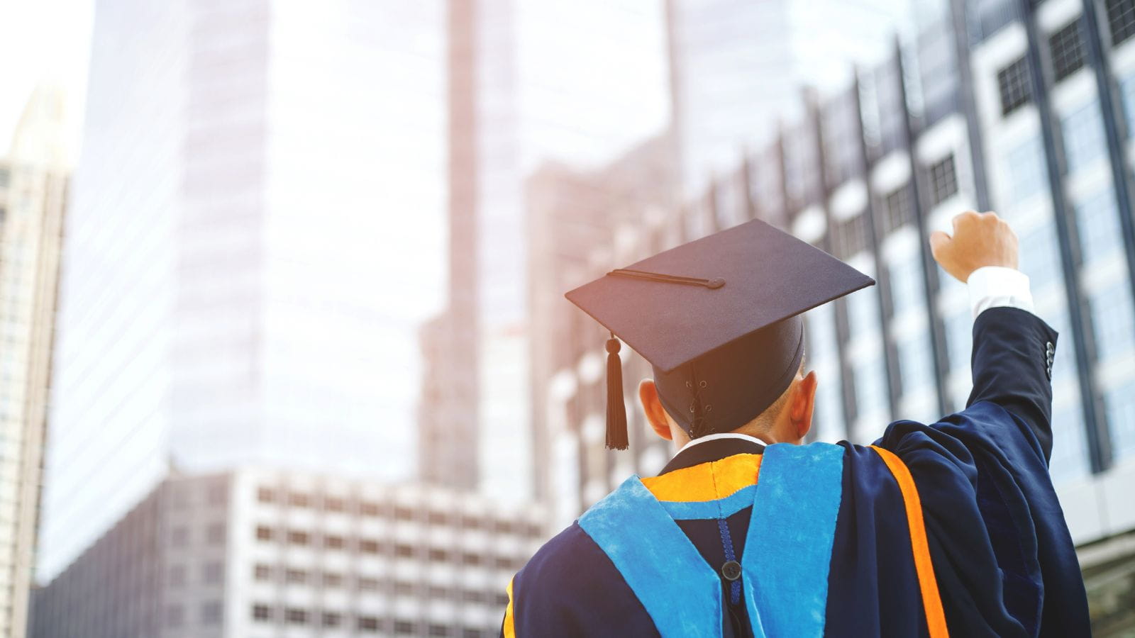 Rear view shot of a young man wearing a graduation cap and gown. He is standing in front of glass office buildings and gesturing triumphantly into the air.