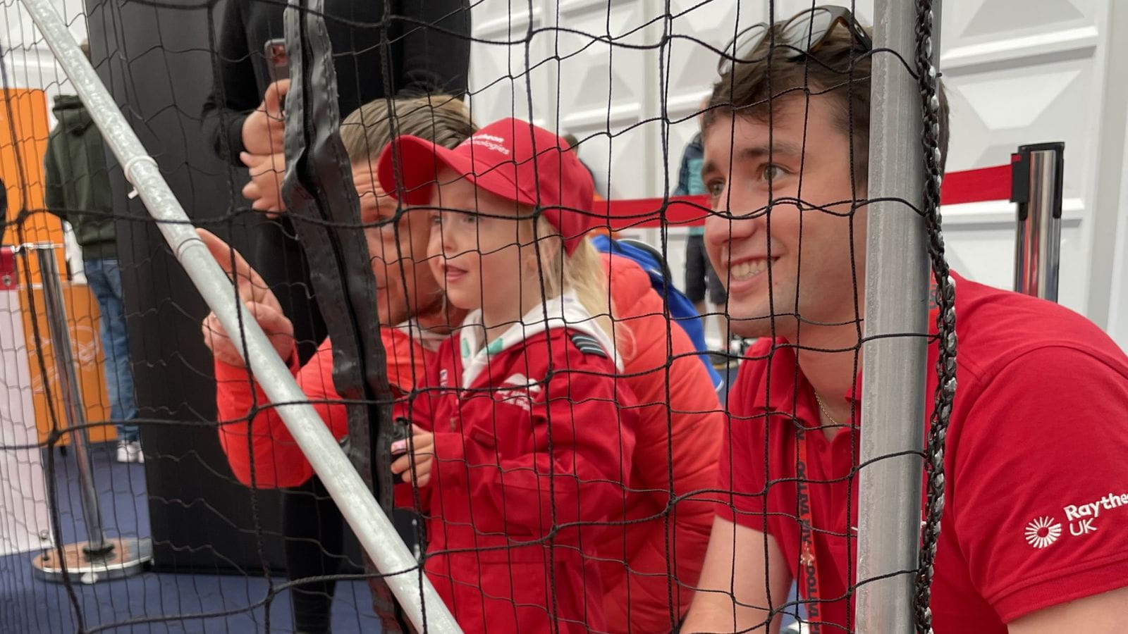 Side profile of a young man wearing a red Raytheon UK T-shirt, with sunglasses on his head. He is kneeling behind a mesh fence, smiling and looking up and to the left. Next to him is a small blonde girl of about three, in a red flight suit and a red Raytheon UK cap. Knelt down on the far side of her is a light-haired man in a red long-sleeve top, looking up and pointing to the top left. 