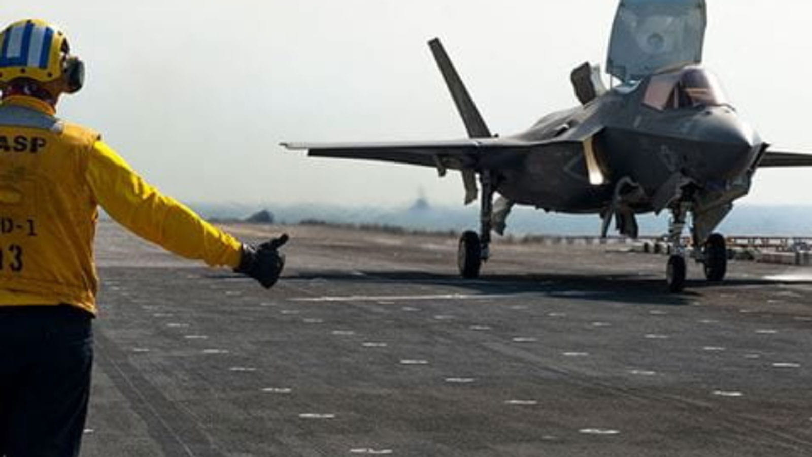 fighter jet taking off from an aircraft carrier, a technician in a yellow suit giving thumbs up