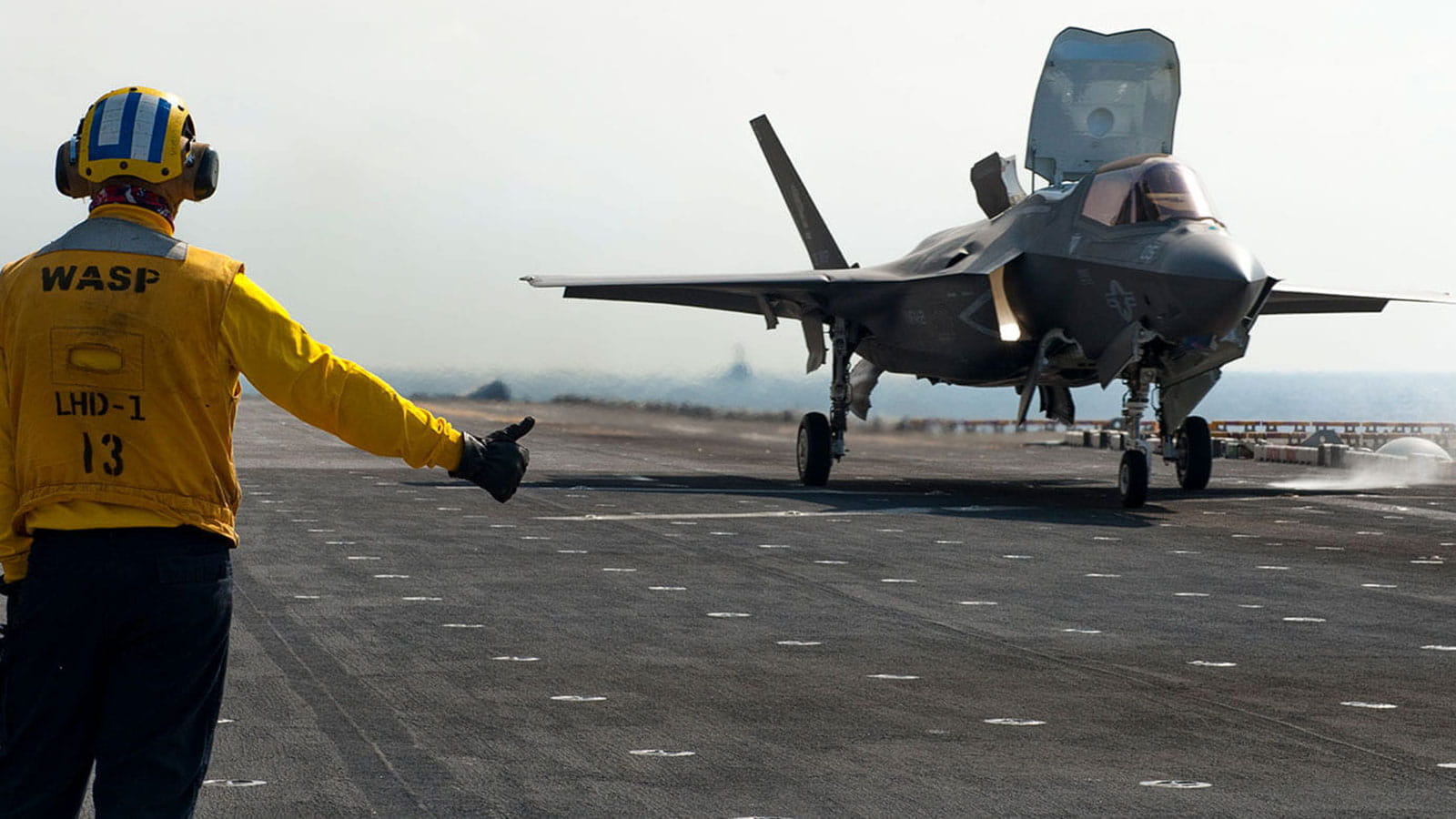 fighter jet taking off from an aircraft carrier, a technician in a yellow suit giving thumbs up