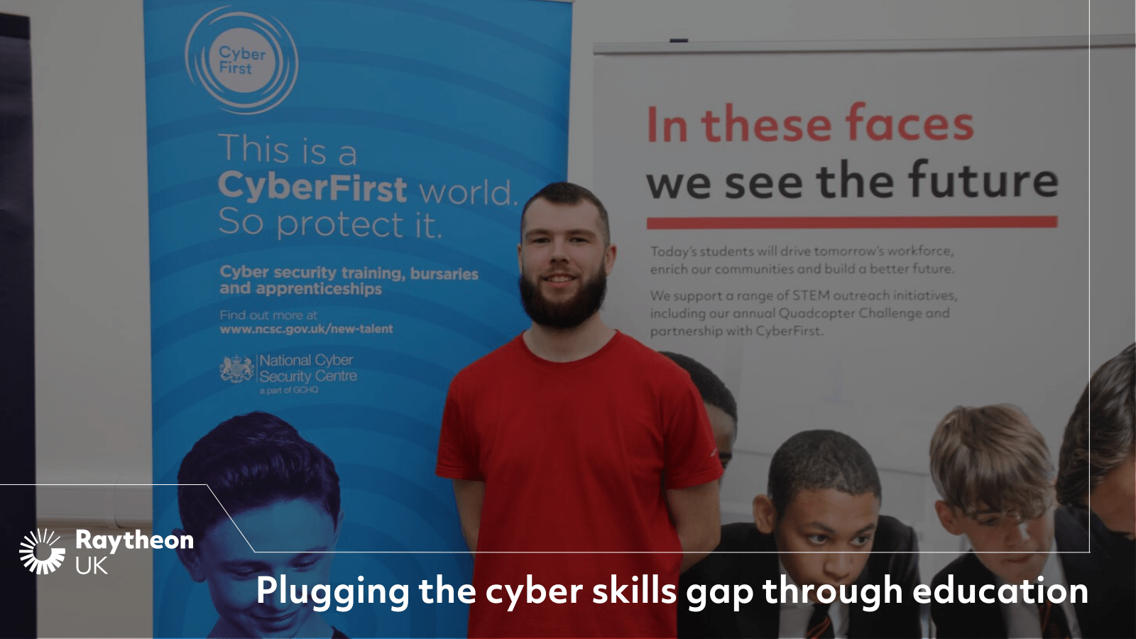 A bearded man in a red t-shirt stands in front of a CyberFirst Schools banner