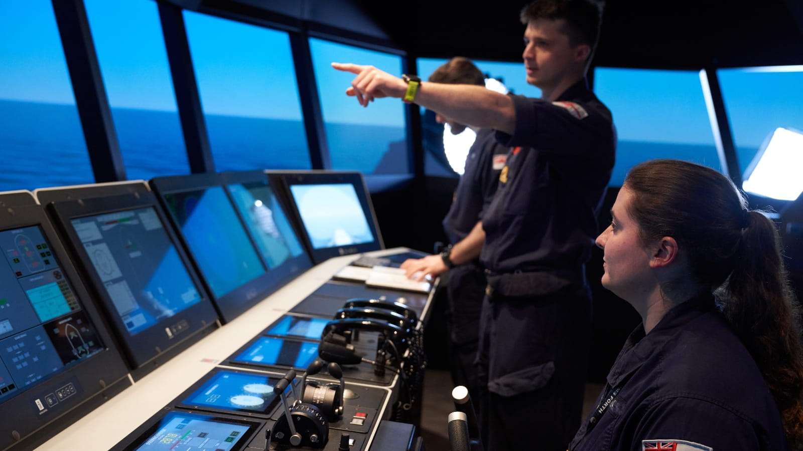 Royal Navy personnel at a ship's control panel. One is steering the vessel, another points out to sea.