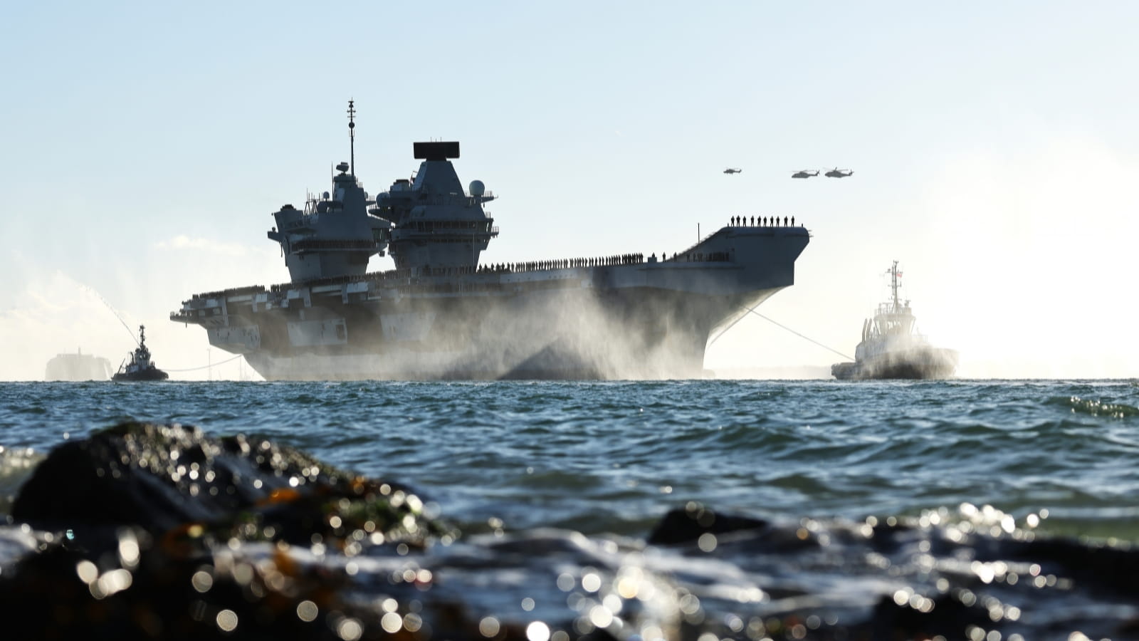 A large navy vessel in the distance, with rocks in the foreground