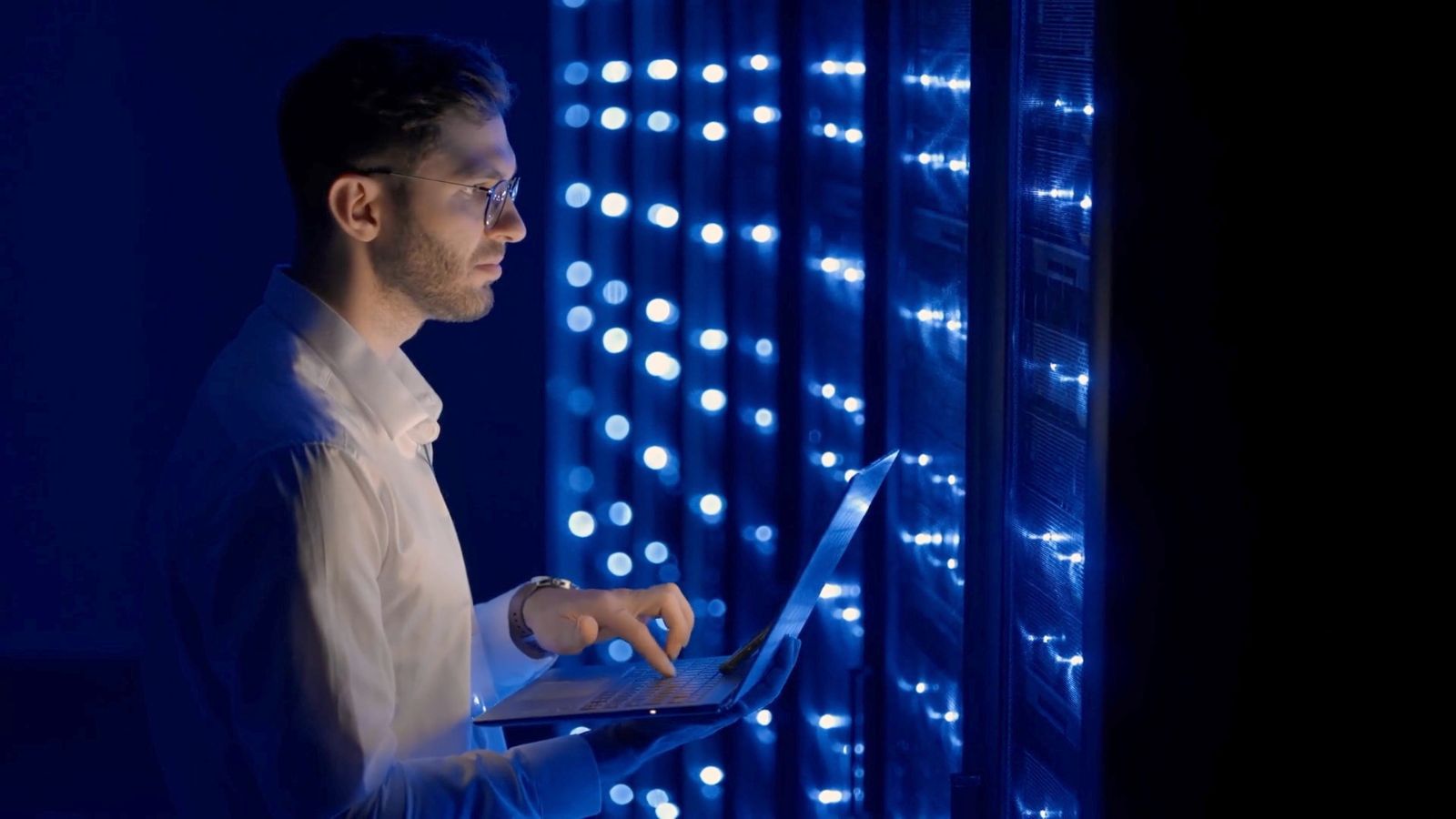 A man stands at a server unit and types on his laptop