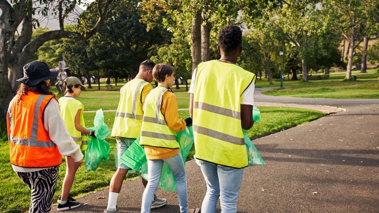 A group of people, facing away from the camera, walk through a park wearing high visibility vests and carrying green litter bags