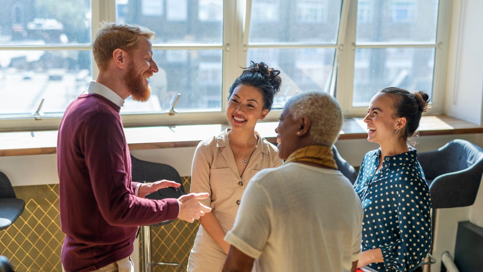 A diverse group of professionals stand in a circle chatting. They are smiling at each other as the sun enters through a large window behind them.