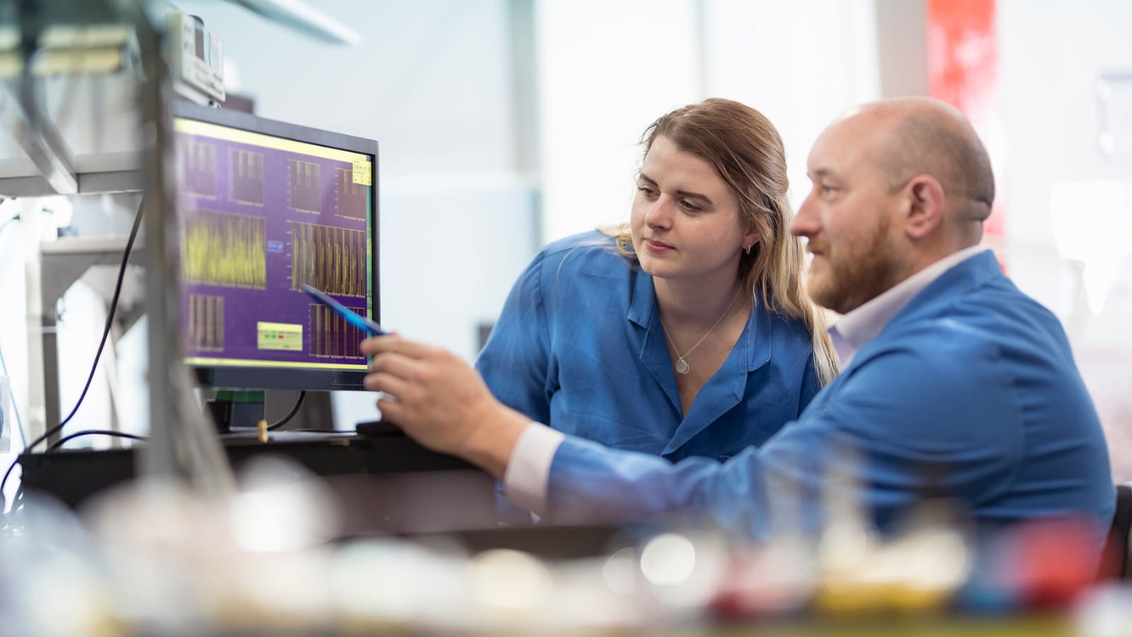 A man and a woman in blue lab coats working at a computer screen