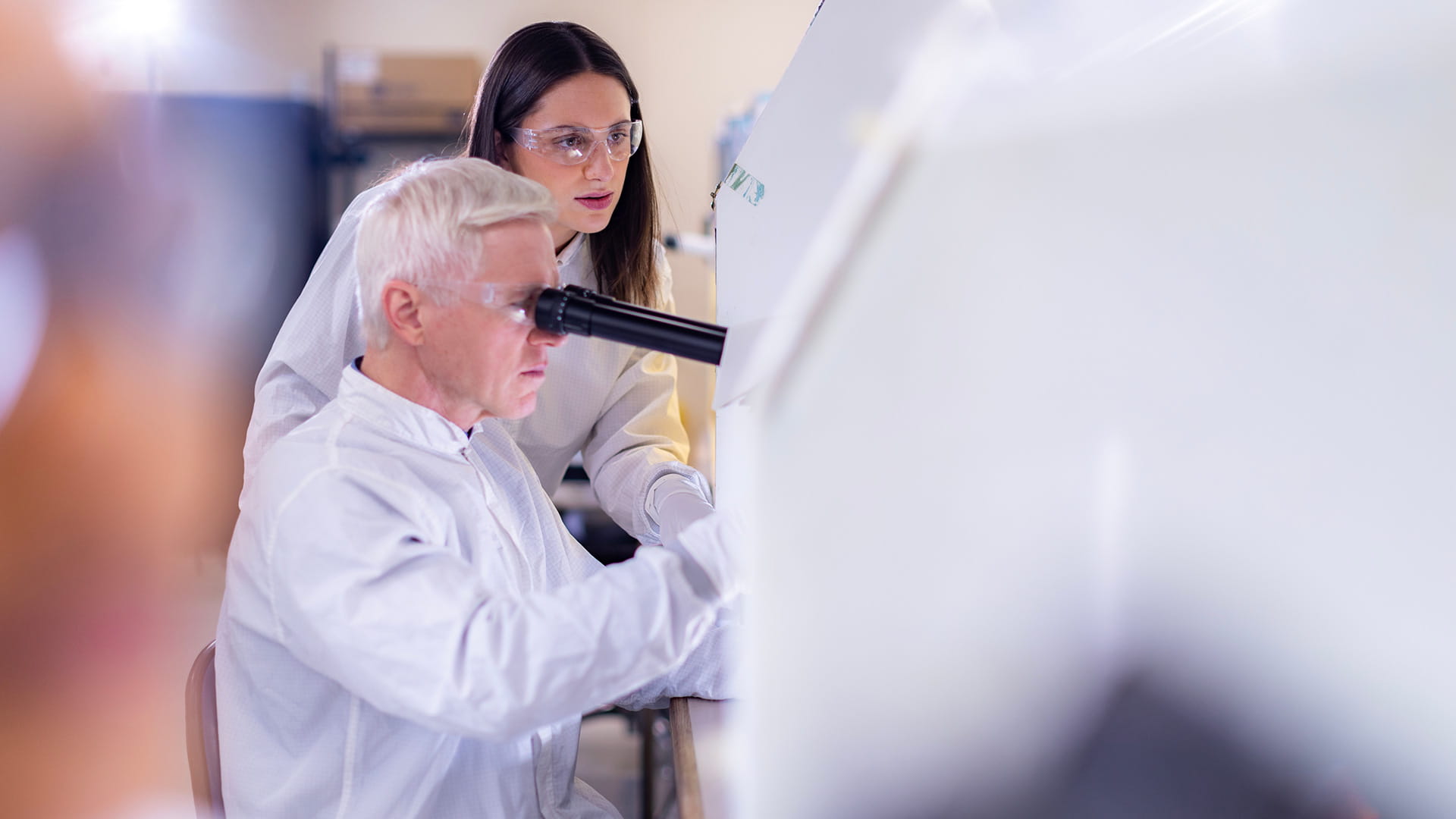 A man peering into a microscope while a woman looks on