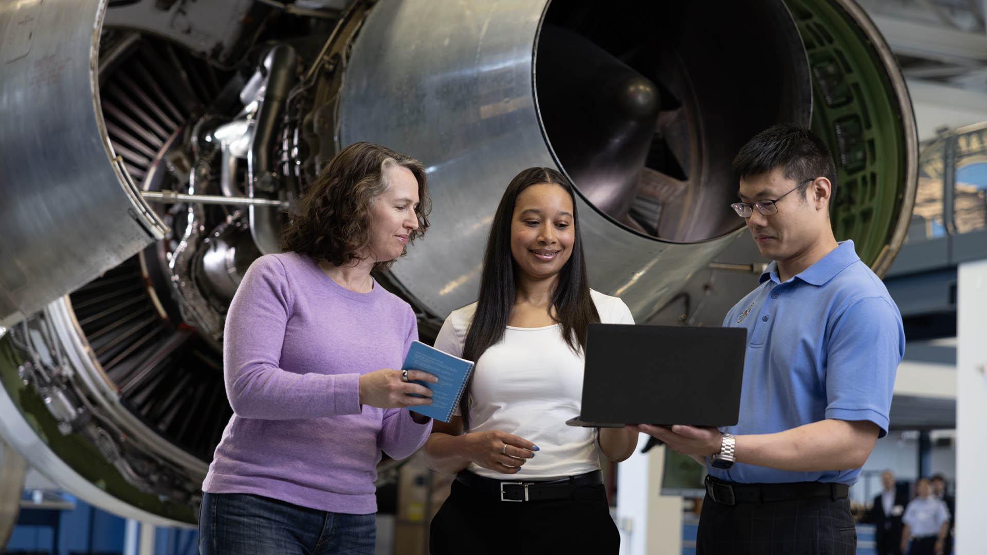 Three people looking at a laptop in front of an aircraft engine