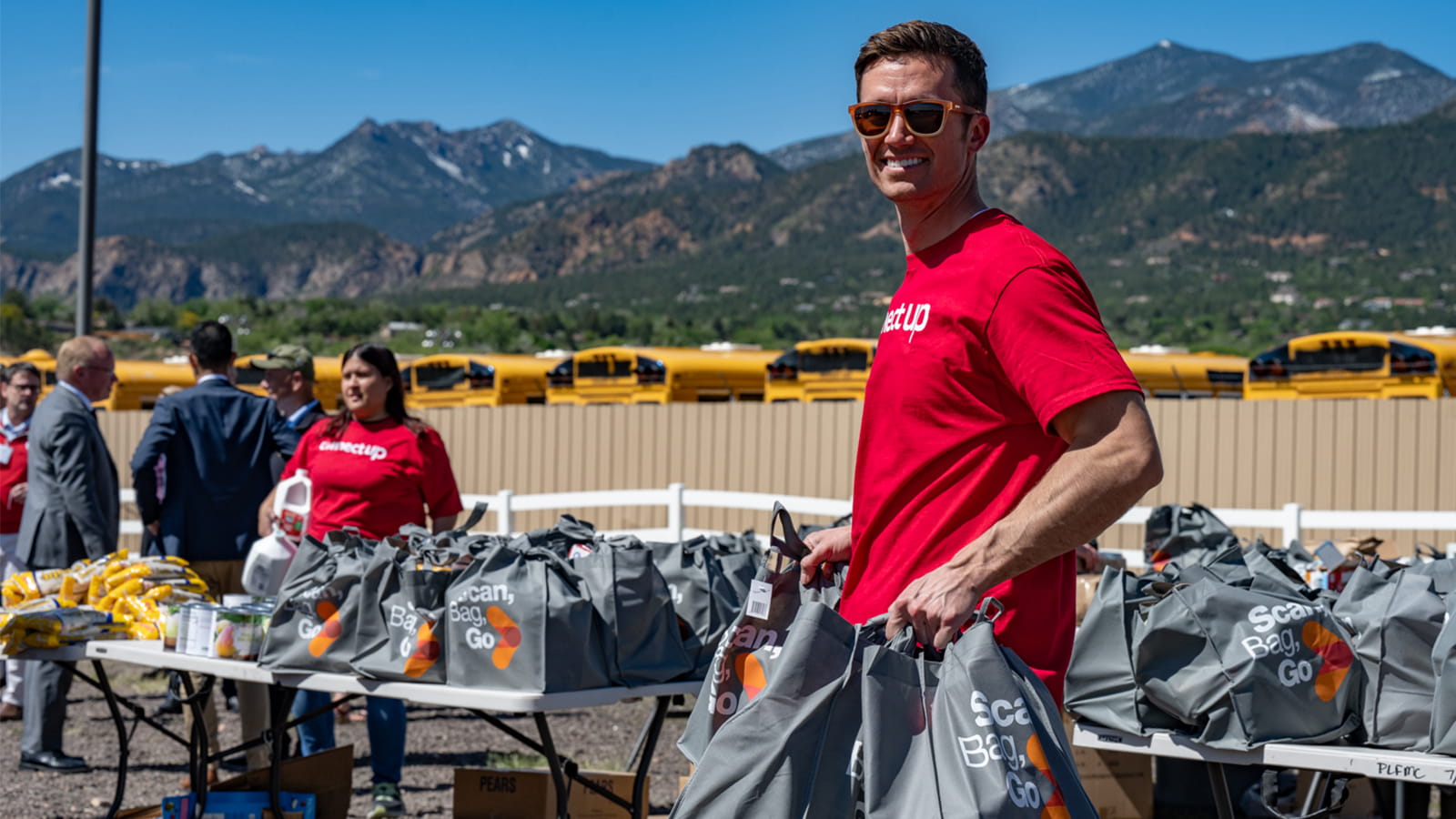 A volunteer, standing against a backdrop of mountains and clear sky, carries two bags of groceries during an event to provide food security for military families in Colorado.