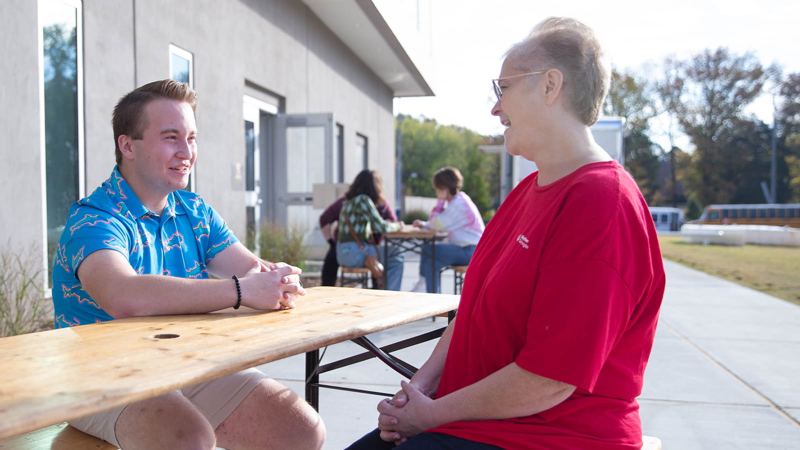 A woman in a red shirt speaks with a young man in a blue polo, sitting across from her at a picnic table.