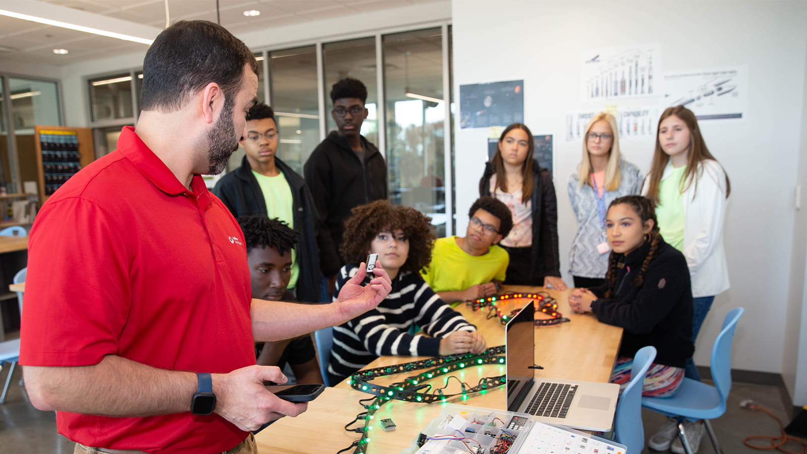 A man in a red shirt shows a microcontroller to a group of nine students gathered around a table in a classroom. A string of green and red LED lights lies on the table, next to a laptop computer and a container of various electronics parts.