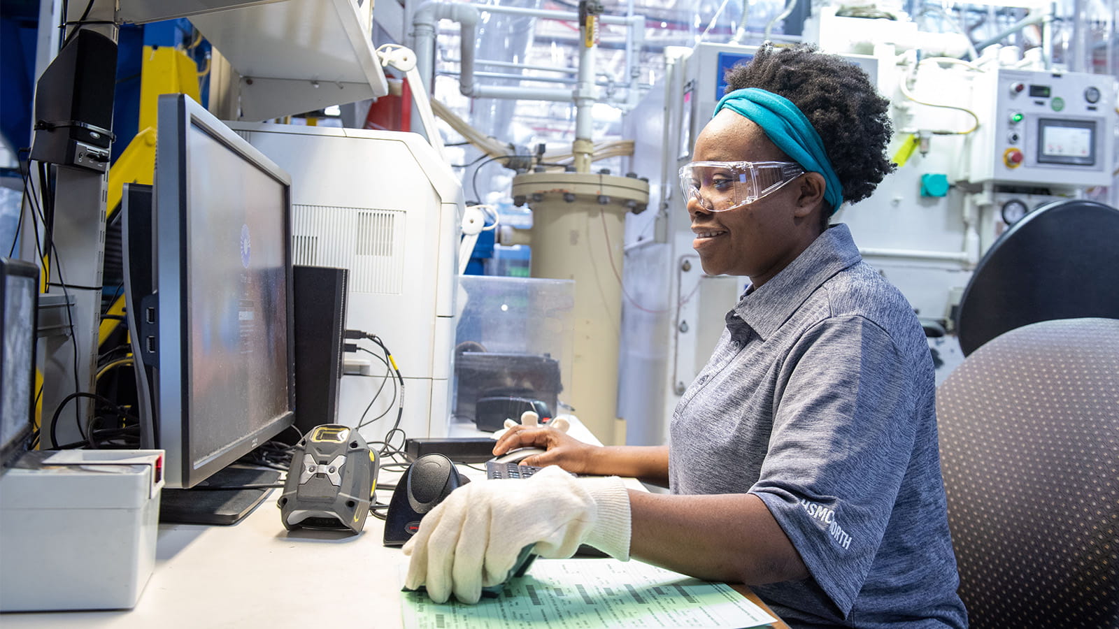 A woman wearing protective eyewear uses a computer mouse while working in a manufacturing facility.