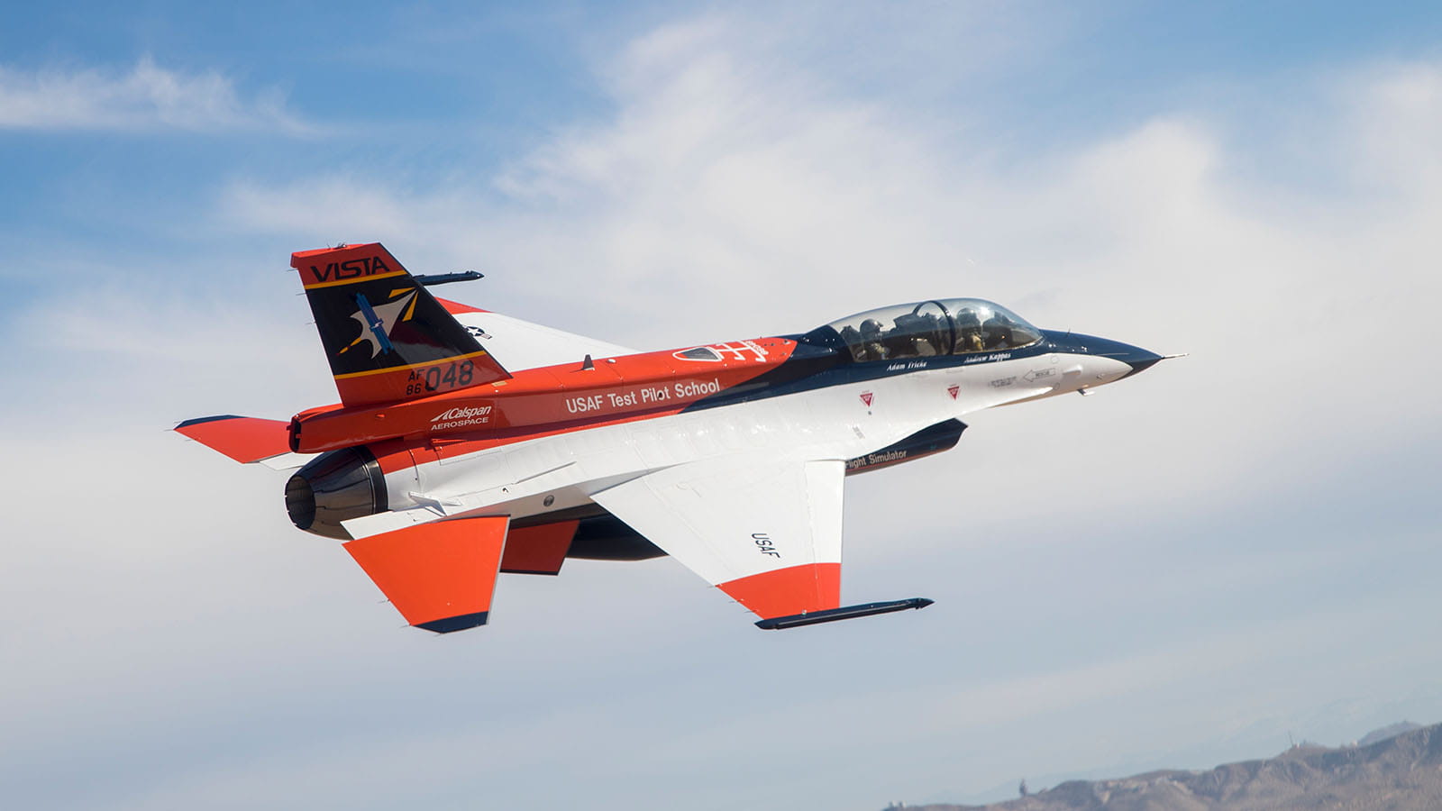 A U.S. Air Force Test Pilot School test aircraft, simliar to an F-16 fighter jet but painted orange, white and blue, flies against a backdrop of blue sky and white clouds.