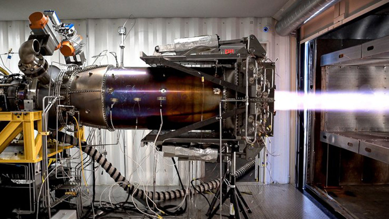 An aircraft engine in a testing rig emits a bright bluish-white exhaust stream.