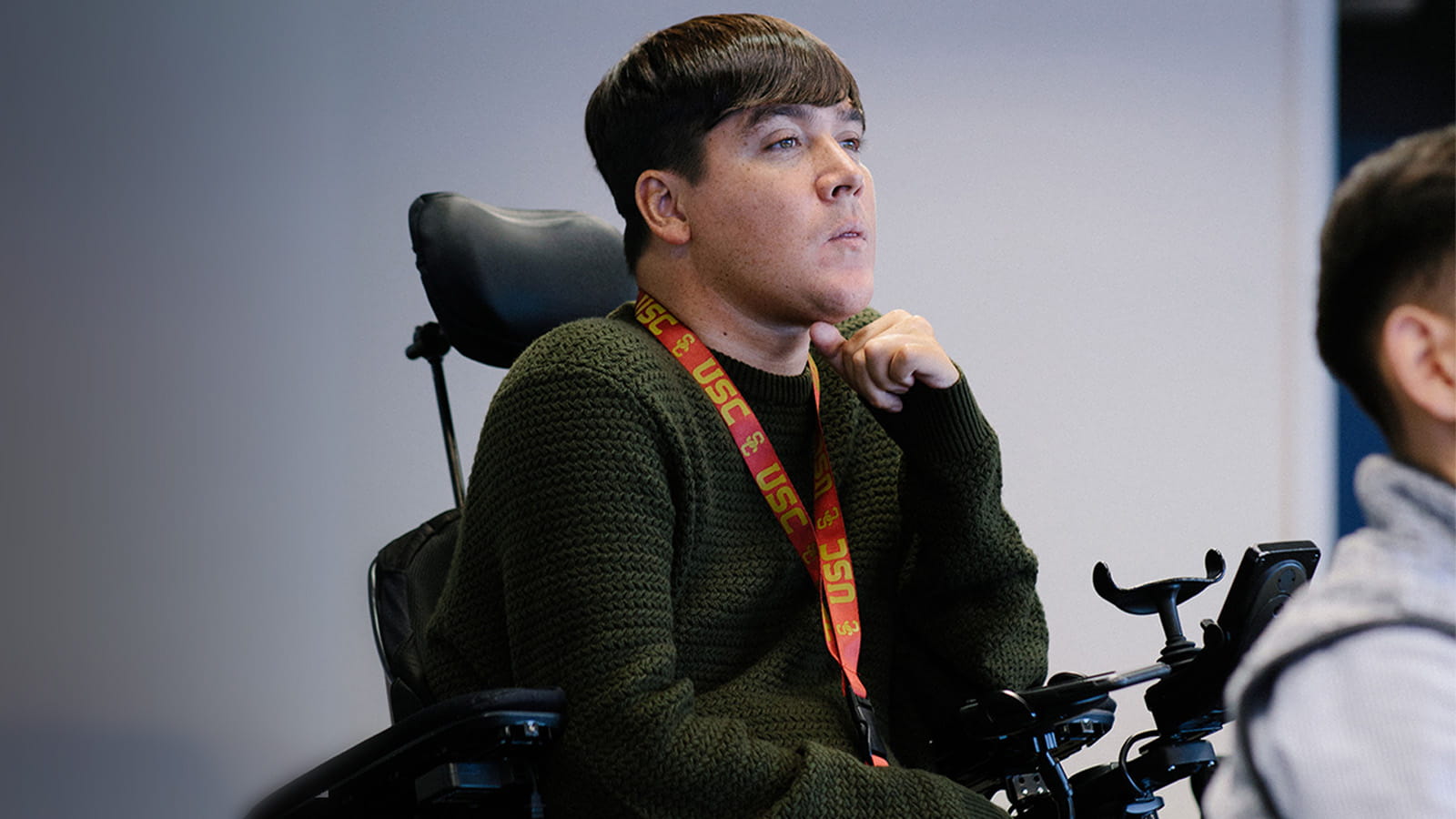 A man with short, dark hair, wearing a green sweater and jeans and sitting in a wheelchair, listens to a presentation in a conference room.