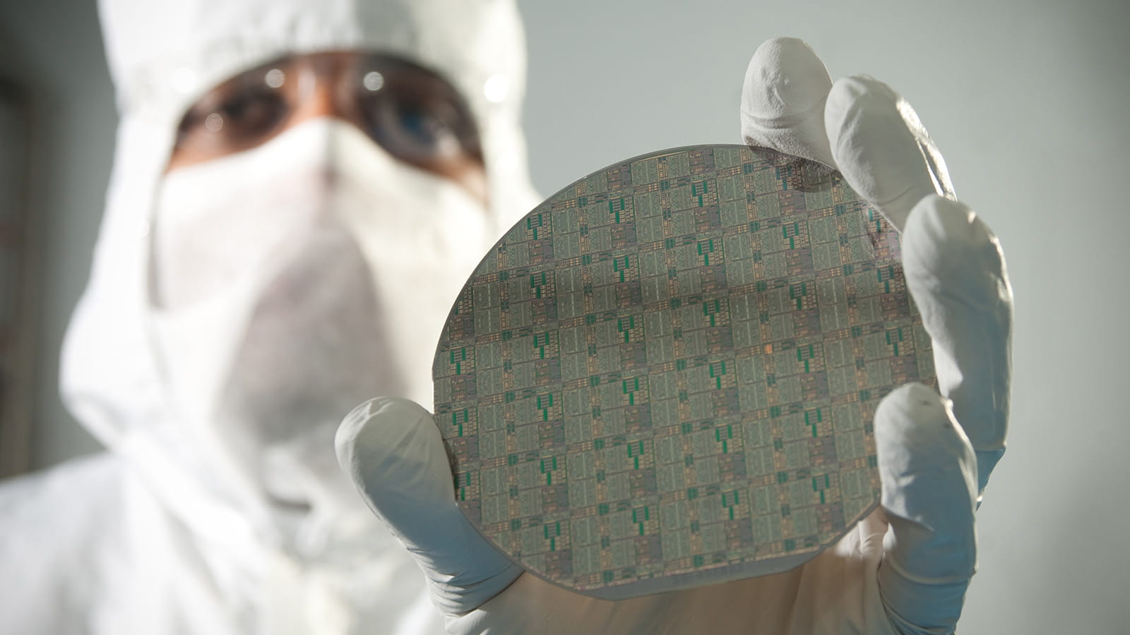 A lab technician wearing a white full-body clean room suit, a face covering and rubber gloves holds up a disc-shaped circuit card with a shiny, reflective surface