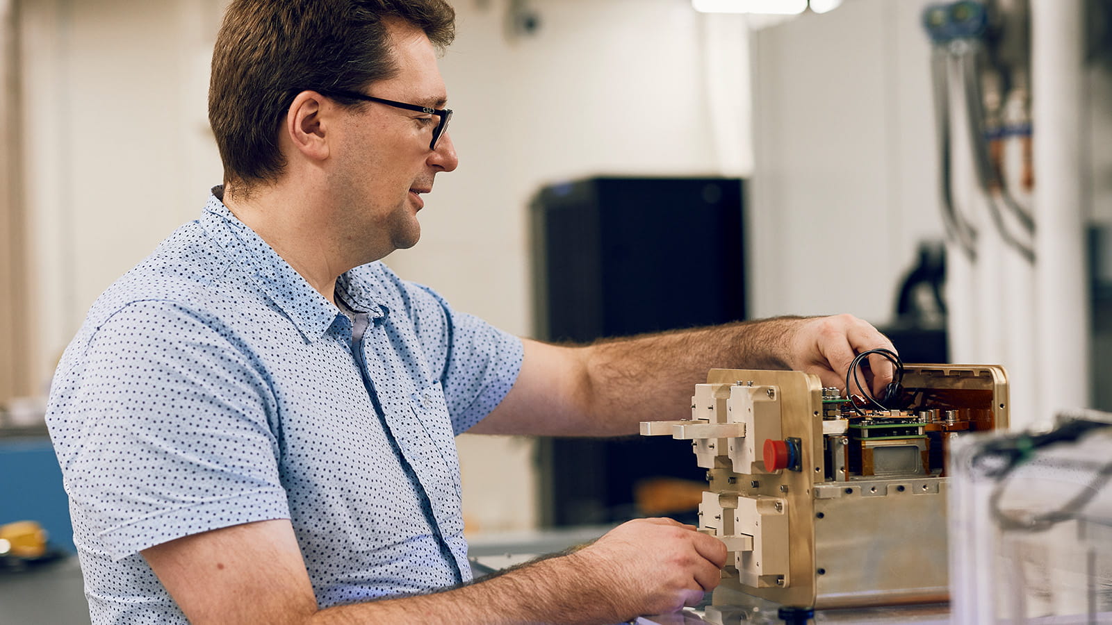 : A man with short brown hair, glasses and a chinstrap beard looks down at a box-shaped circuit breaker that rests on a see-through work surface. He places his left hand on copper-insulated electrical wiring and uses his right to hold a beige-colored metallic rod.