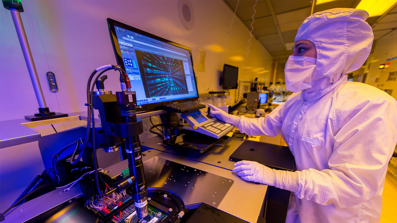 A lab worker wearing an all-white clean room suit, gloves and a mask uses a joystick to operate a microscope while looking at a computer monitor.