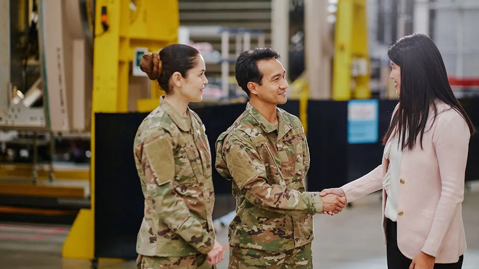 two people in military uniforms are greeted by a woman in a shop floor environment.