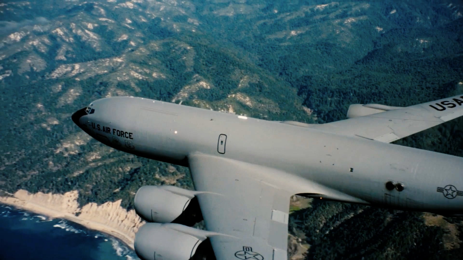 A large military airplane flies over green trees