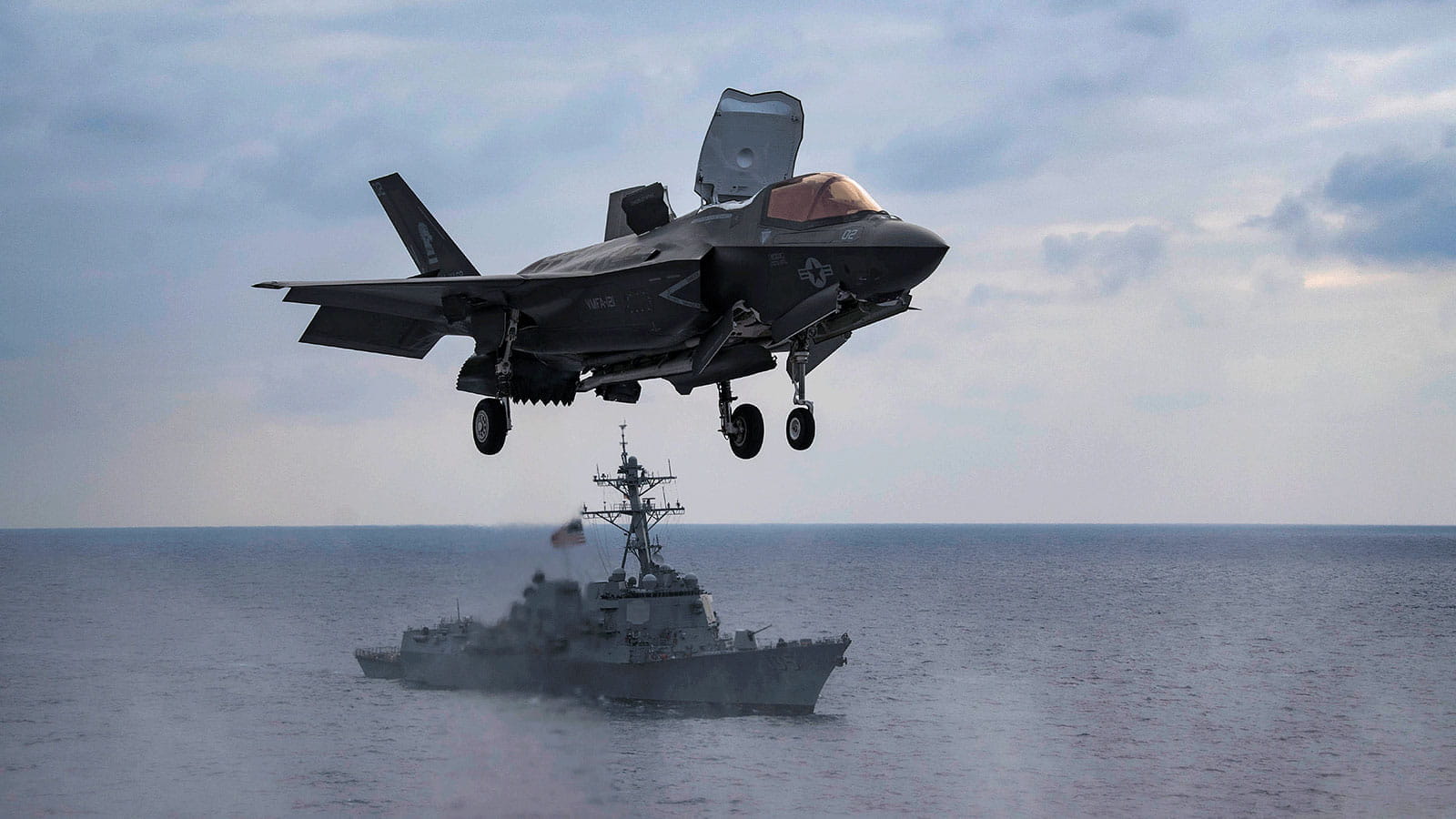 An F35 taking off or landing at sea with a battleship behind it.
