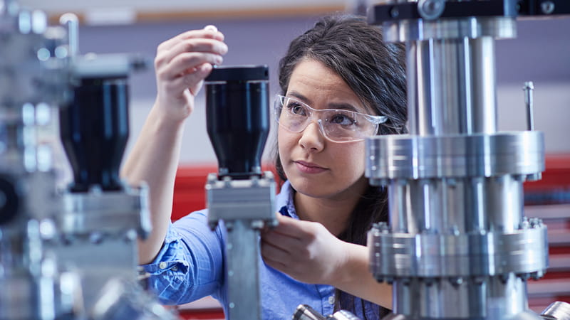 A scientist wearing safety glasses is adding a substance to stainless steel apparatus.