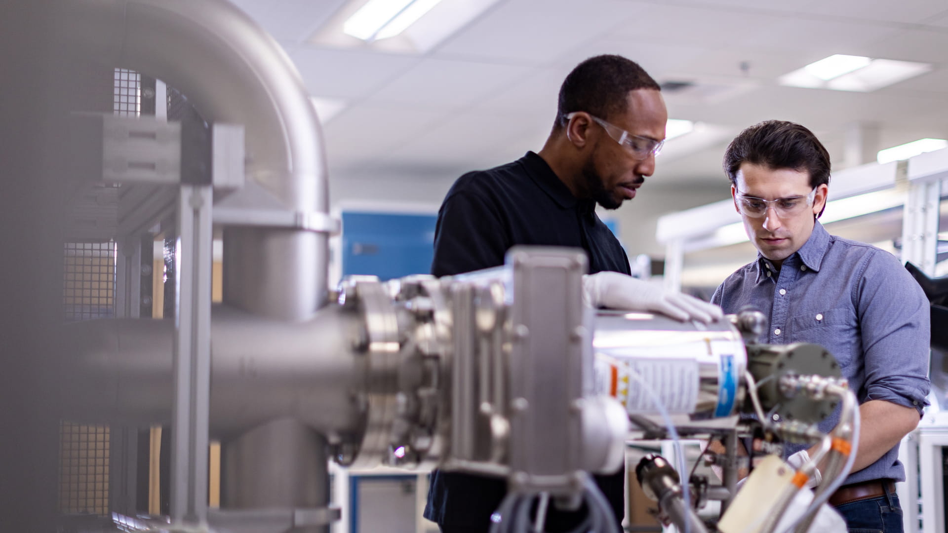 Two adult males both wearing safety glasses and protective gloves working in a brightly lit lab.