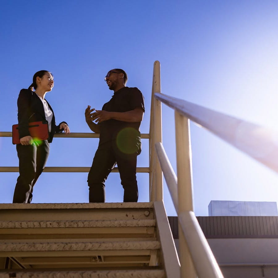 Adult male and adult female having a conversation at the top of a staircase outside on a clear day. 