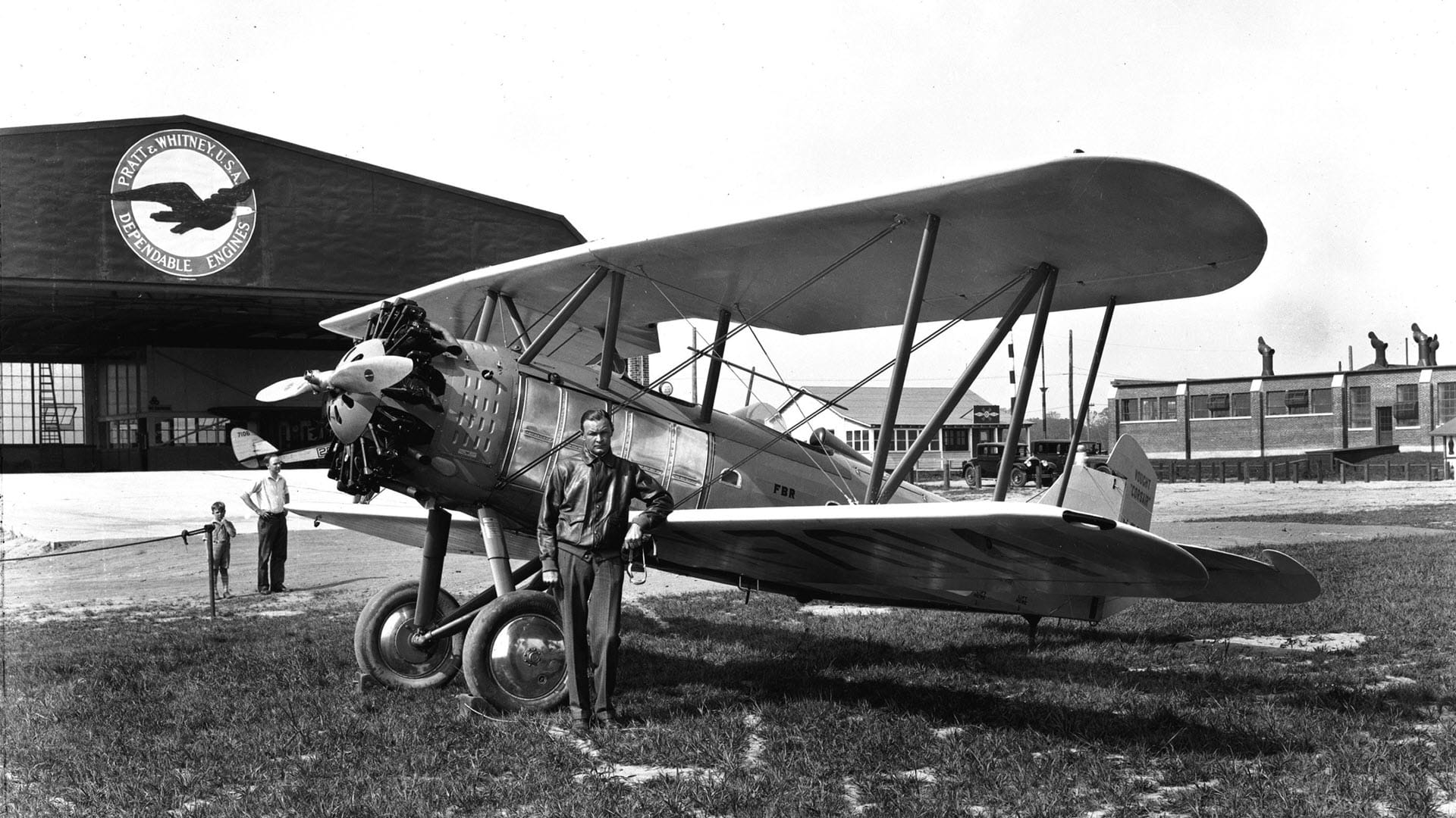 black and white photo of a man in front of a biplane and a hangar is in the background
