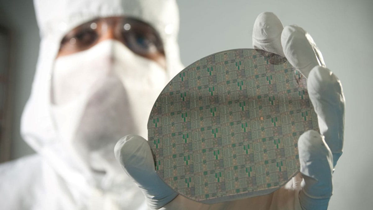 A lab technician wearing a clean suit holds a wafer up to the camera