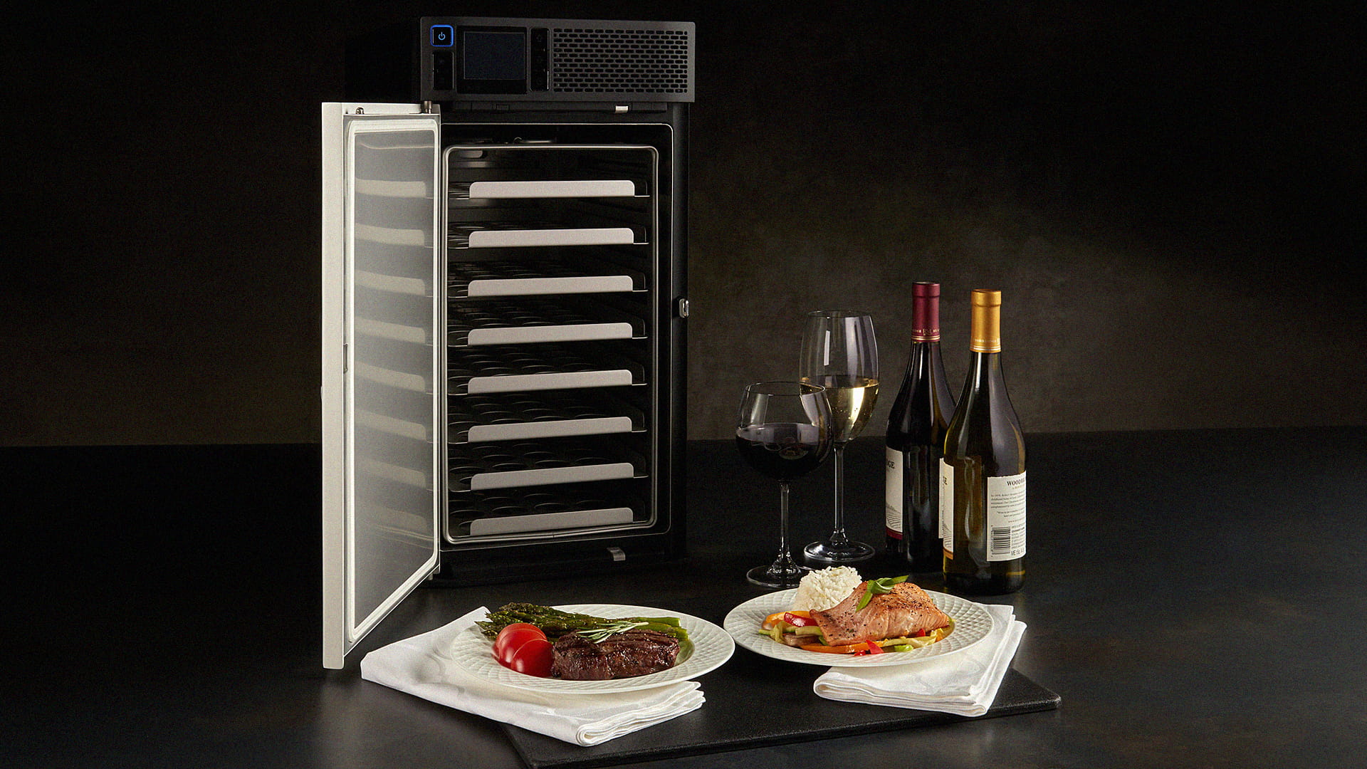A small steam oven, two plates of food, two bottles of wine, and two wine glasses