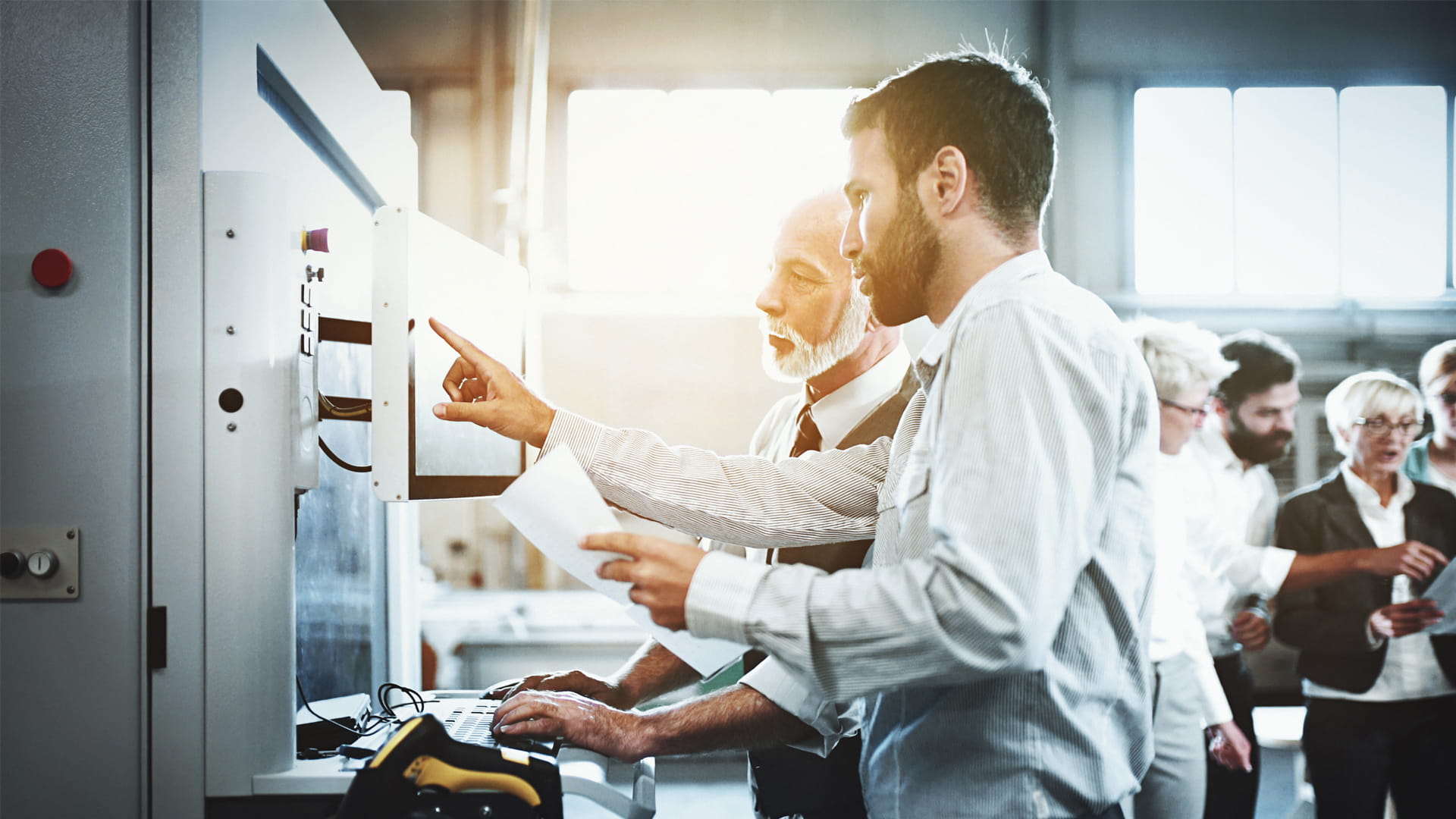 Closeup of senior manager and a mid 30's coworker looking at certain printed data and standing in front of a CNC cutting machine at a wood production facility. There are some of their colleagues in the background. Image by gilaxia