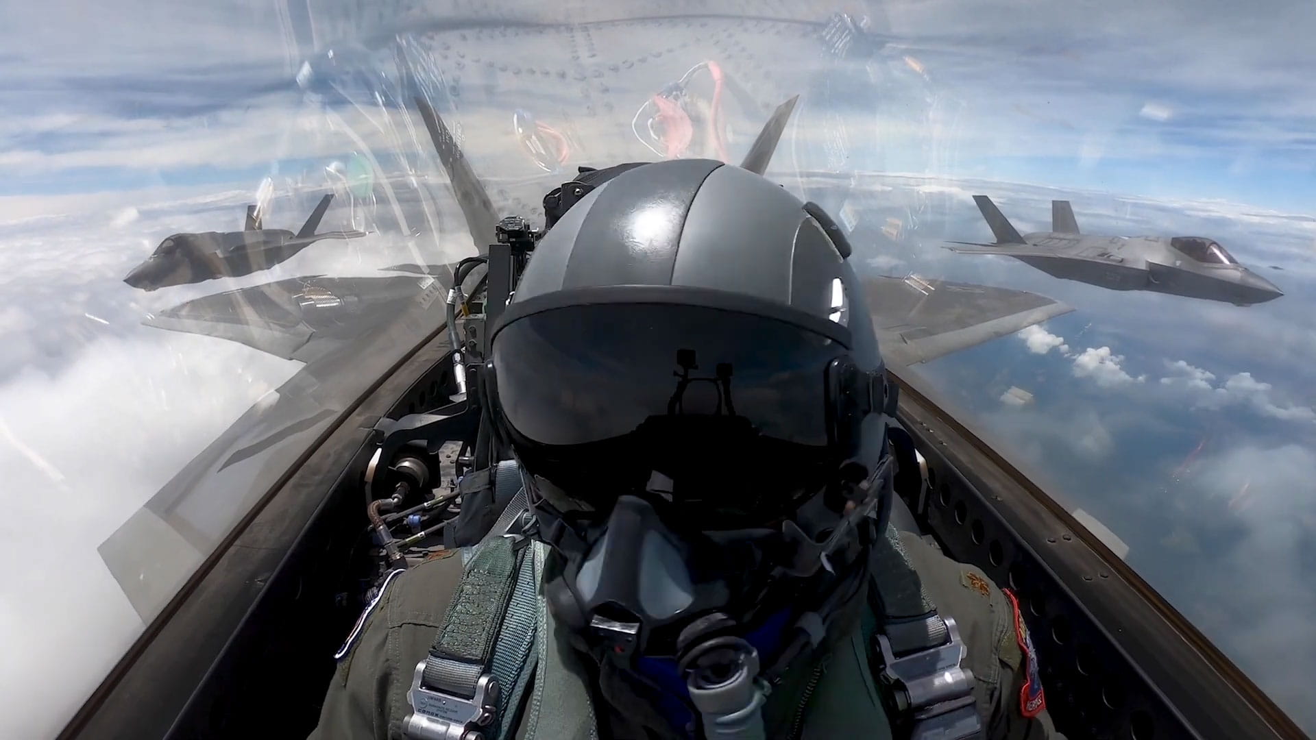 A fighter pilot in the cockpit of a fighter jet with two fighter jets visible out the cockpit window