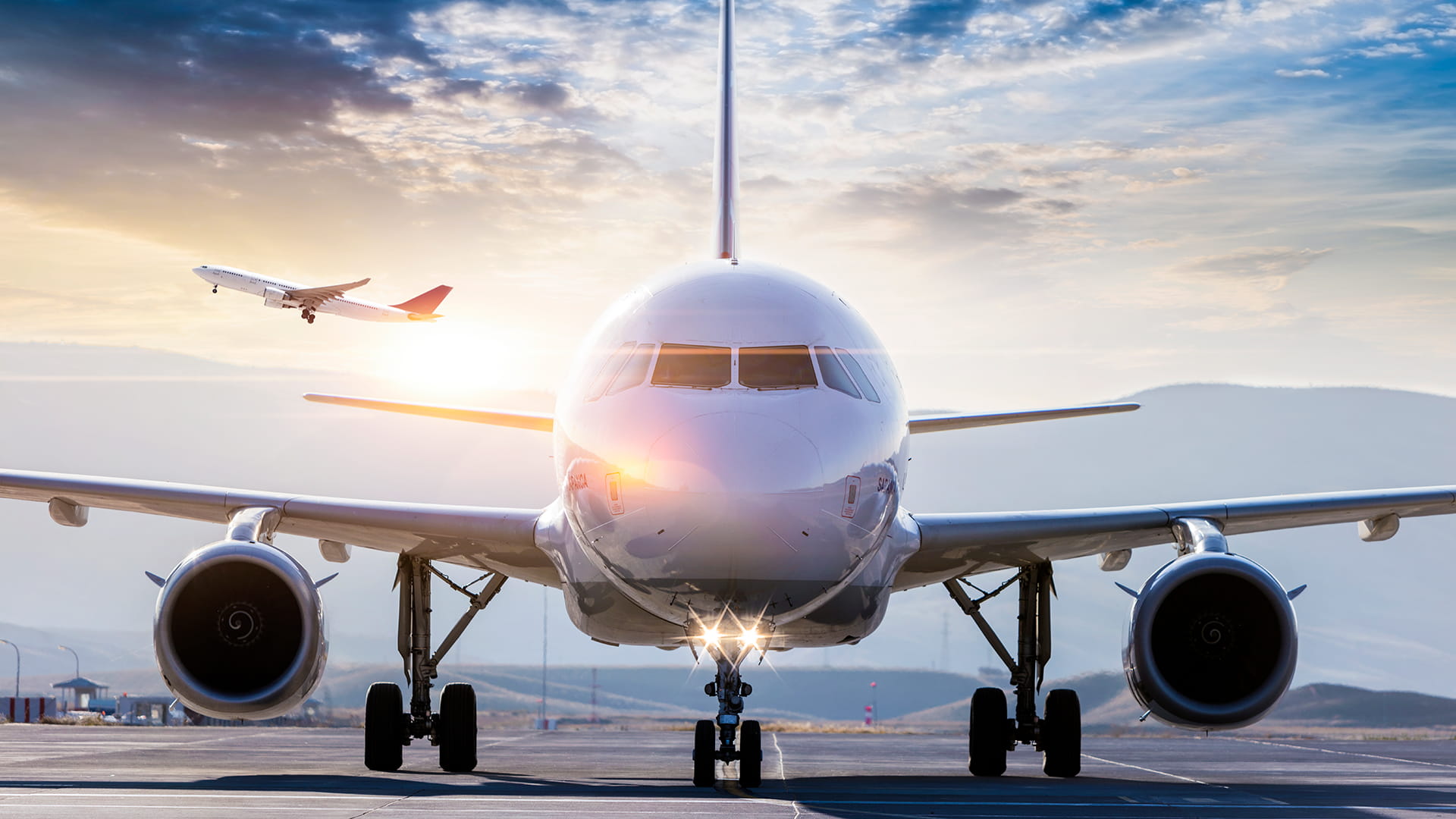 An aircraft sits on the runway while another aircraft takes off in the background