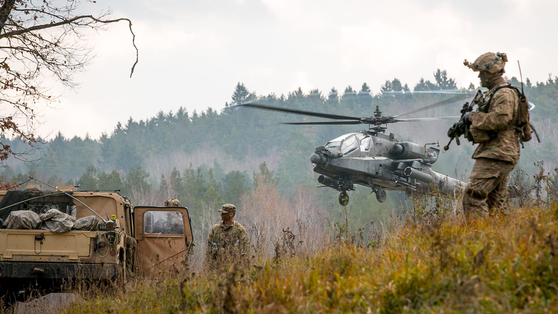 Three warfighters in the field with tactical gear by a Humvee with an Apache helicopter hovering in the background