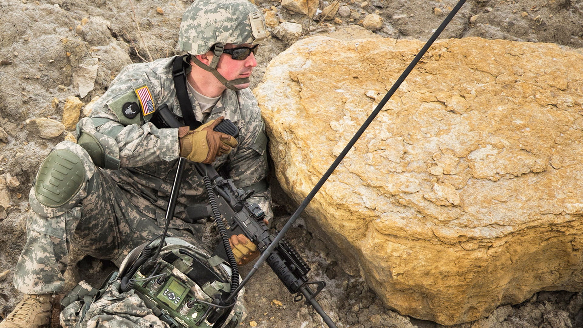 Warfighter in uniform with field communications gear crouching behind large rock