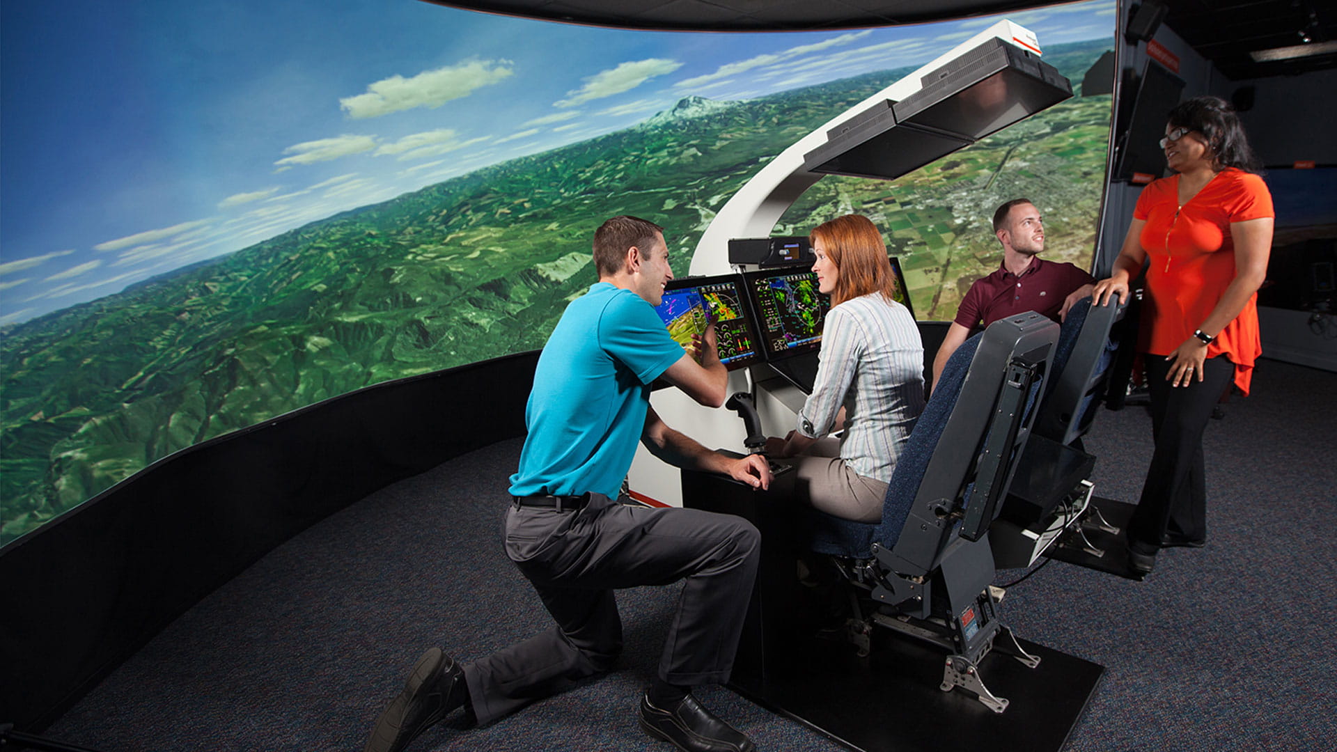 Instructor pointing at monitor screens with operator sitting in seat in front of large-screen simulator