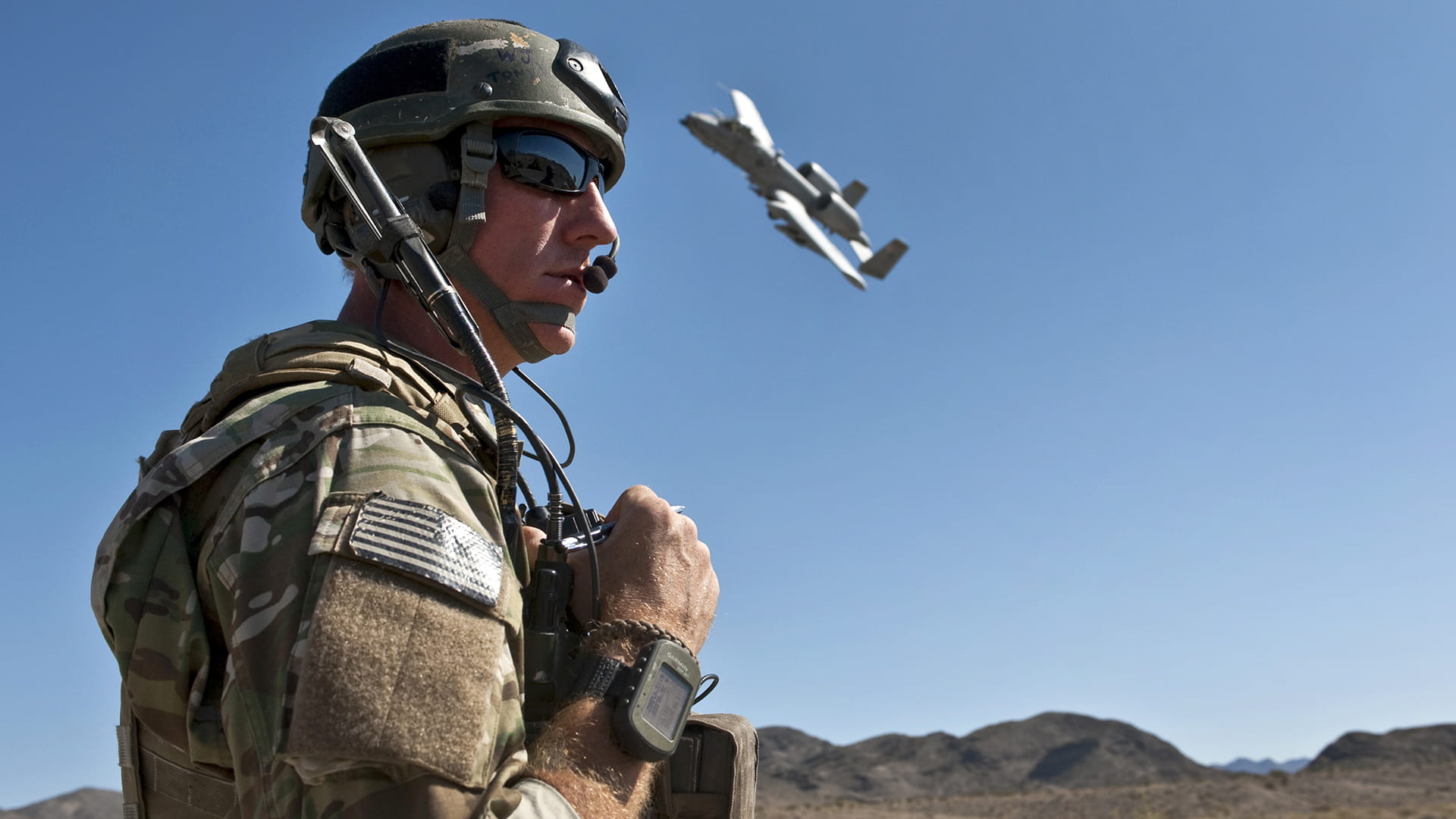 Photo of warfighter in uniform in the foreground with a military plane in flight in the sky behind him