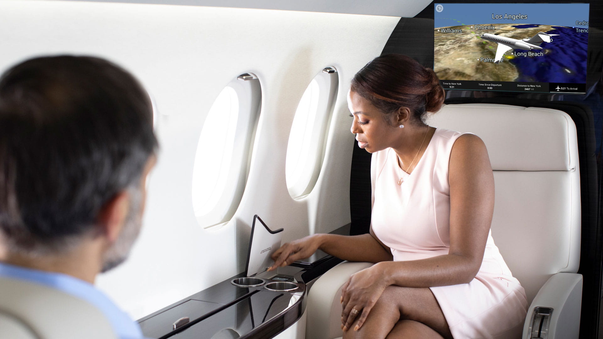 A man and woman in a business jet cabin. A moving map display appears on a large television behind them.