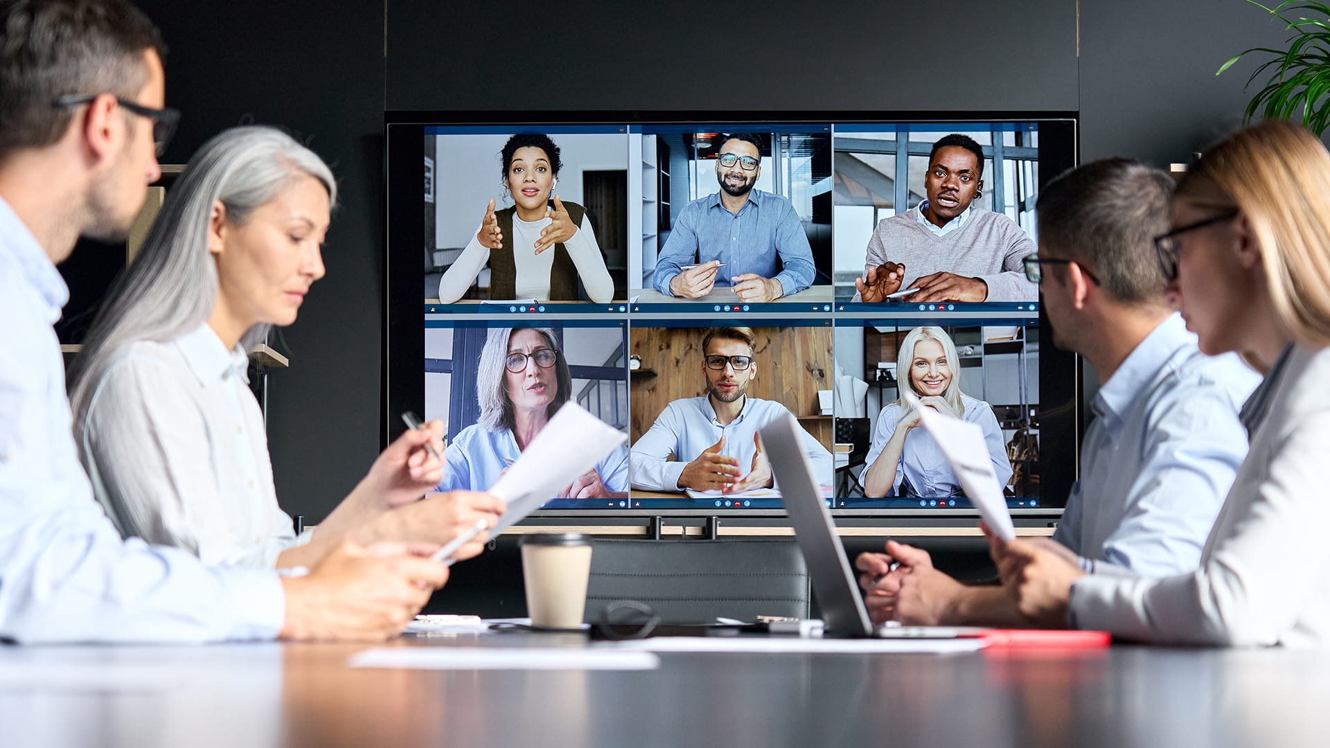 Global corporation online videoconference in meeting room with diverse people sitting in modern office and multicultural multiethnic colleagues on big screen monitor.