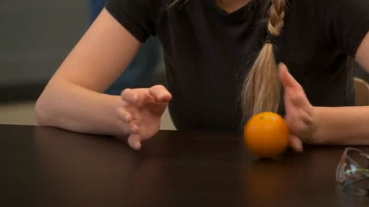 A woman sitting at a table rolling an orange back and forth between her hands