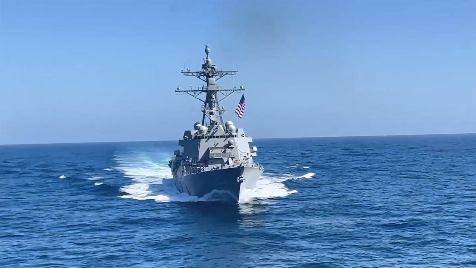A U.S. Navy destroyer ship flying a U.S. flag cuts through blue ocean waters against a clear blue daytime sky