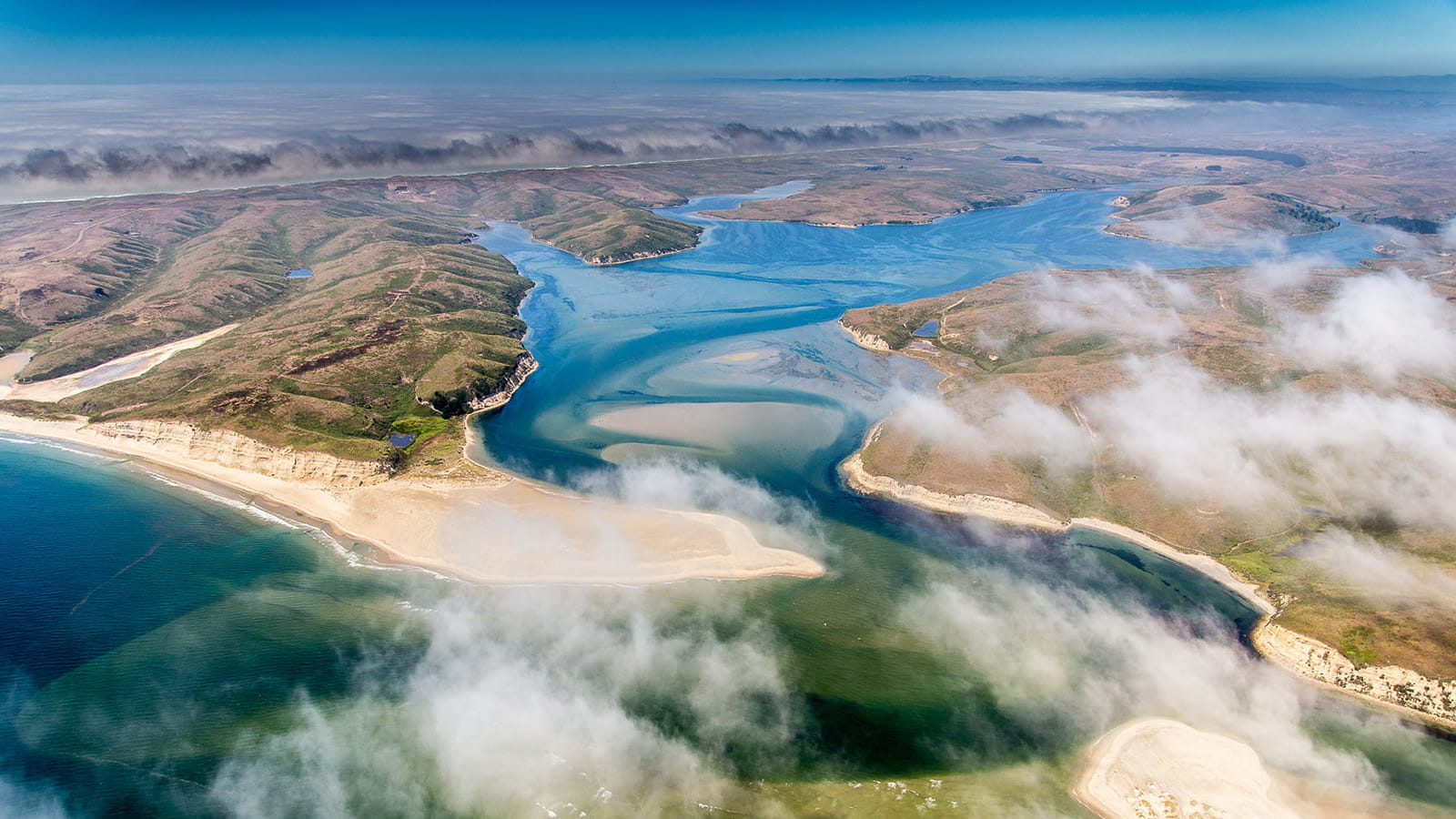 An aerial view of a coastal region in shows blue-green water flowing along a beach and inland, into valleys separated by low-rising greenish brown hills. There are clouds in the foreground.